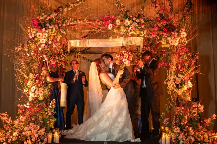 Sharon y Josh bajo la jupá cubierta de flores en tonos rosa y naranja