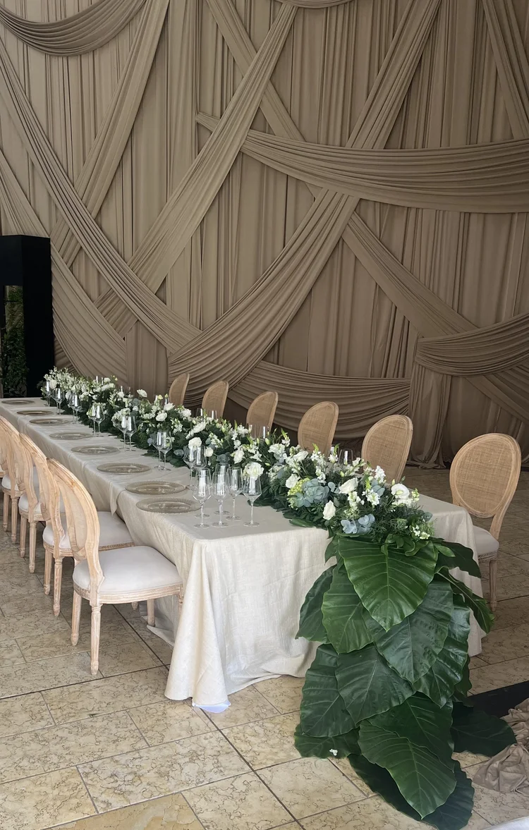 Shabat de André: mesa larga con sillas de ratán, flores blancas y pared de drapeados