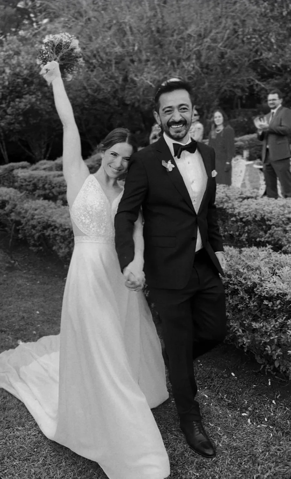 Paloma y Julio recién casados caminan por el jardín de la finca, ella levanta el ramo (blanco y negro)