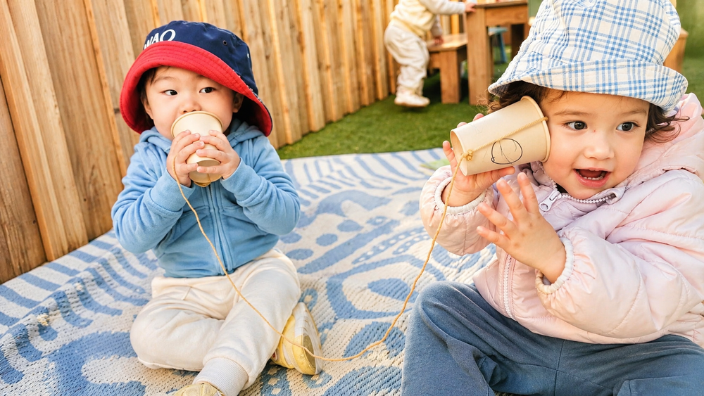 Two toddlers communicating with handmade cup telephones outdoors.