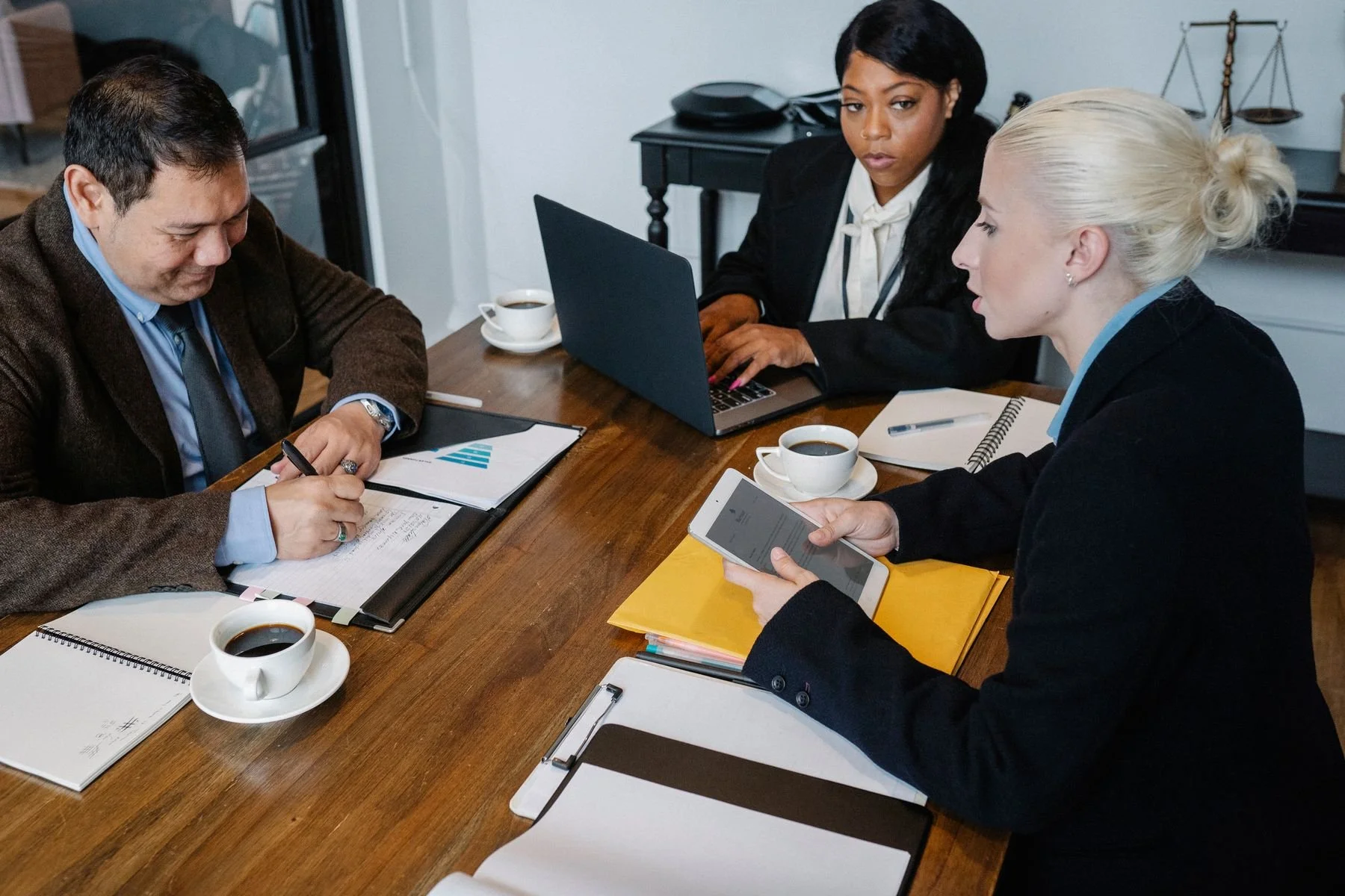 Three professionals considering documents, a laptop and a tablet around a meeting table