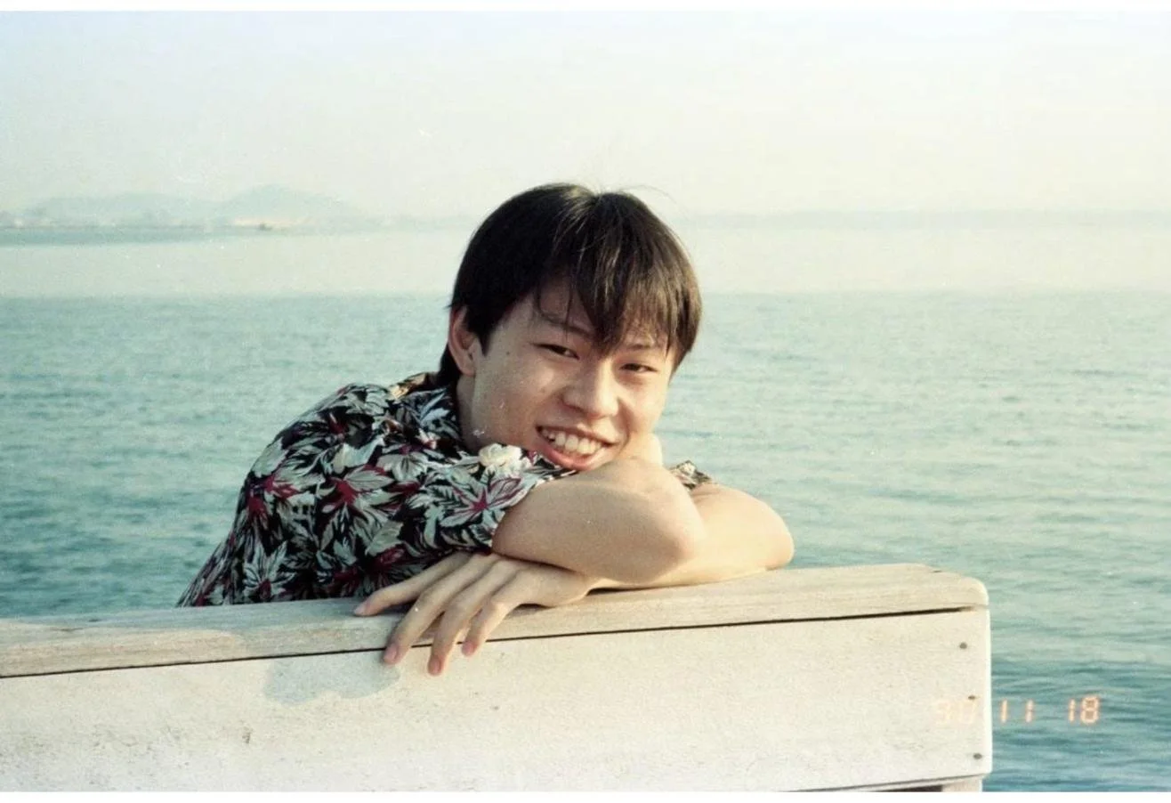 Dr. Rushapol Sdawat as a teenager, smiling and leaning on a wooden rail by the sea, November 1990