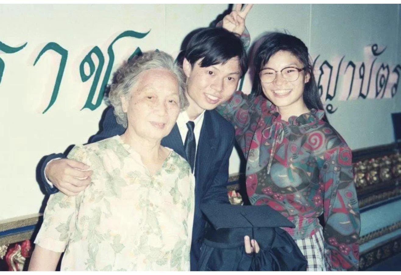 Dr. Rushapol Sdawat in a dark suit at a graduation ceremony, holding a mortarboard, with family beside him
