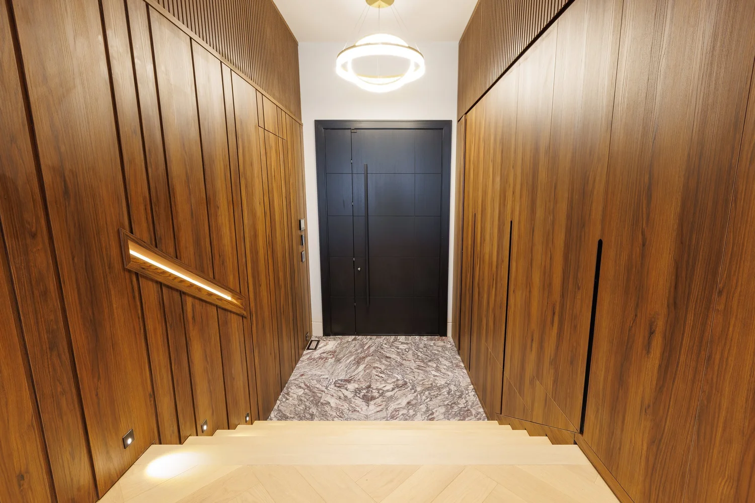 Foyer lined in full-height walnut panelling with concealed closets, an integrated handrail, and a black entry door.