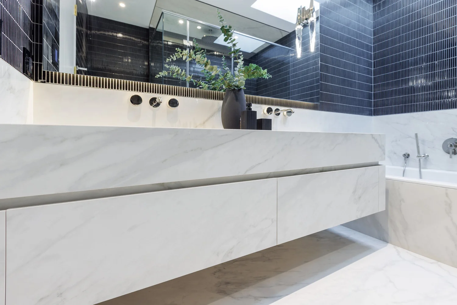 Floating marble double vanity with wall-mounted taps, dark tiled walls, and an adjacent marble bathtub.