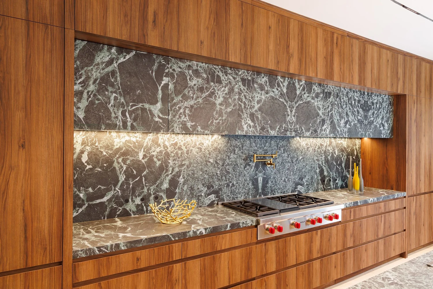 Cooking wall with a green marble backsplash, integrated marble hood, and brass pot filler set into walnut cabinetry.