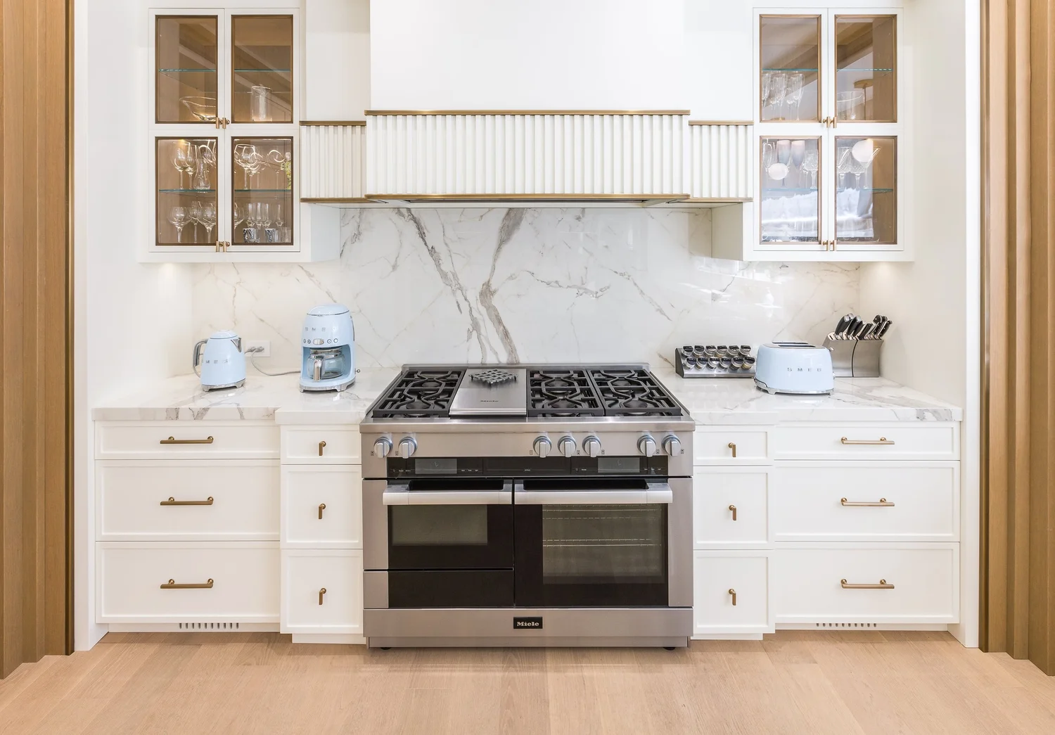 Range wall with a fluted hood, marble backsplash, and glass-front upper cabinets.