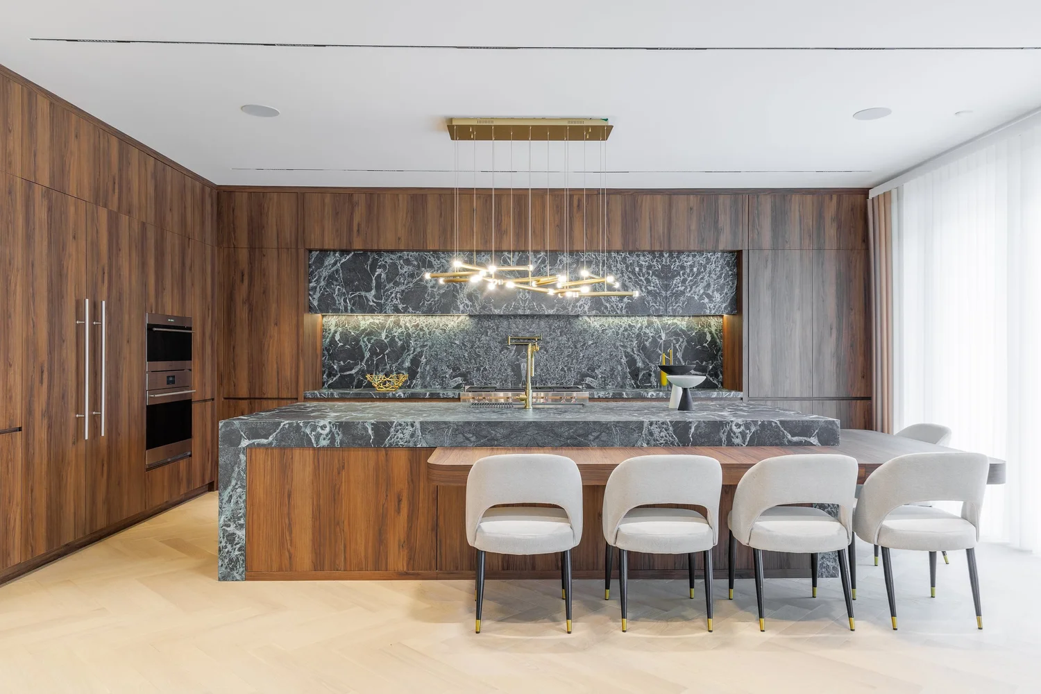 Frontal view of the green marble island with its integrated walnut dining table beneath a brass linear pendant.