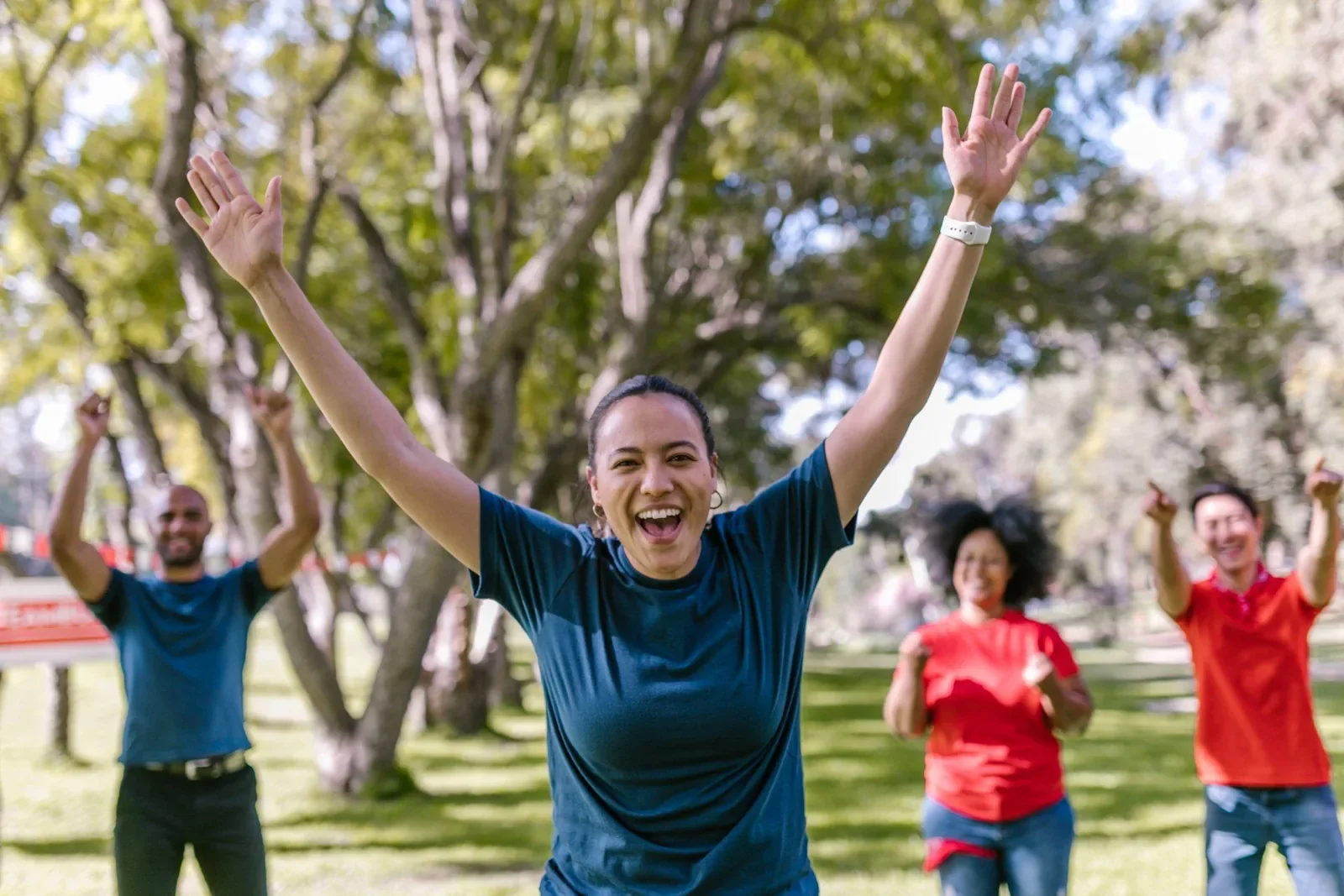 A woman in a park laughing with both arms raised, friends cheering behind her