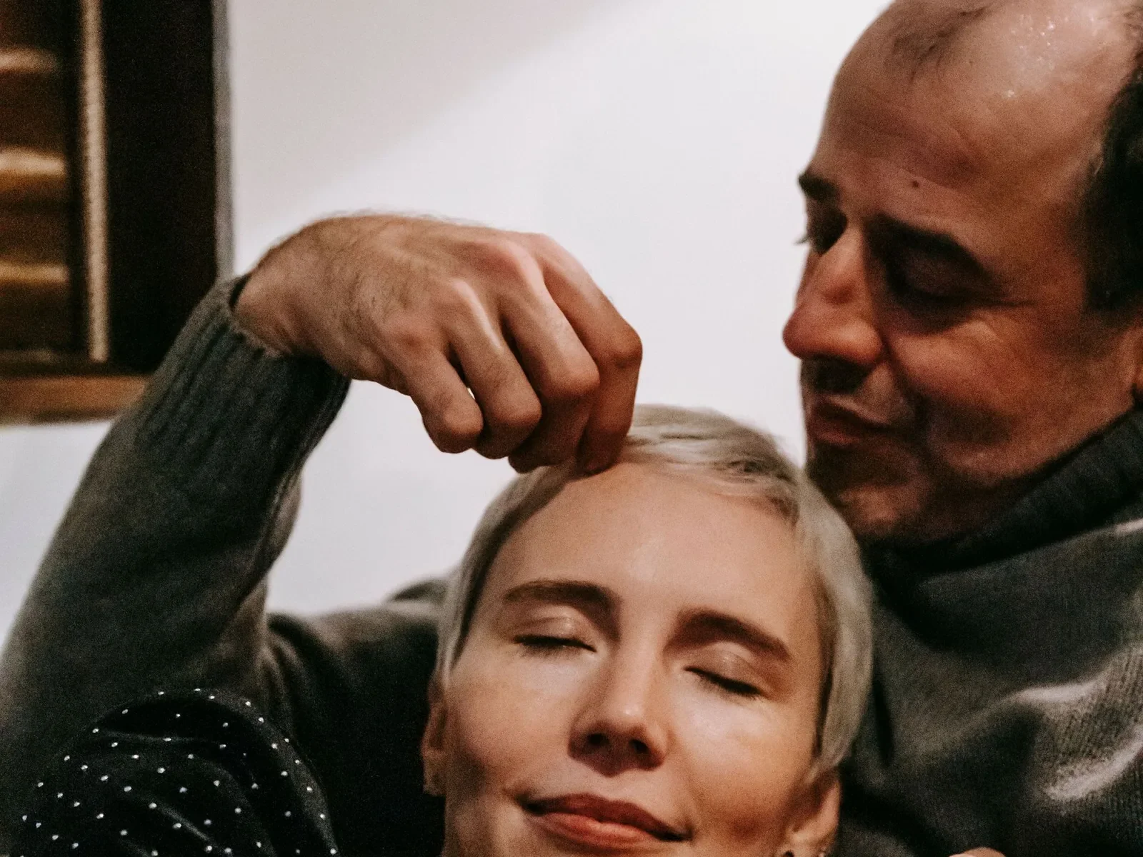 An older couple in a quiet embrace at home, her eyes closed as he gently touches her hair