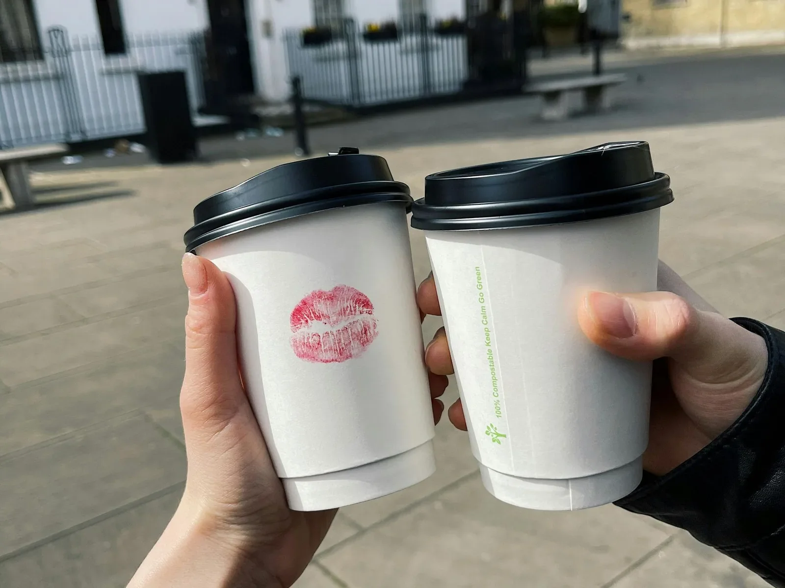 Two hands raising takeaway coffee cups in a toast on a city street, one cup marked with a lipstick print