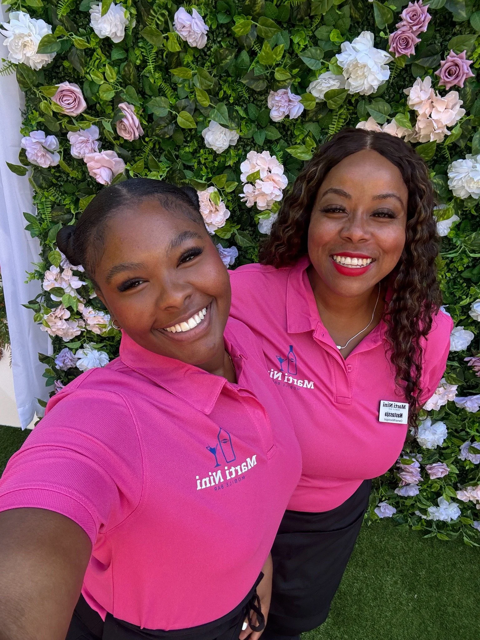 Two Marti NiNi bartenders smiling in pink uniforms in front of a floral wall