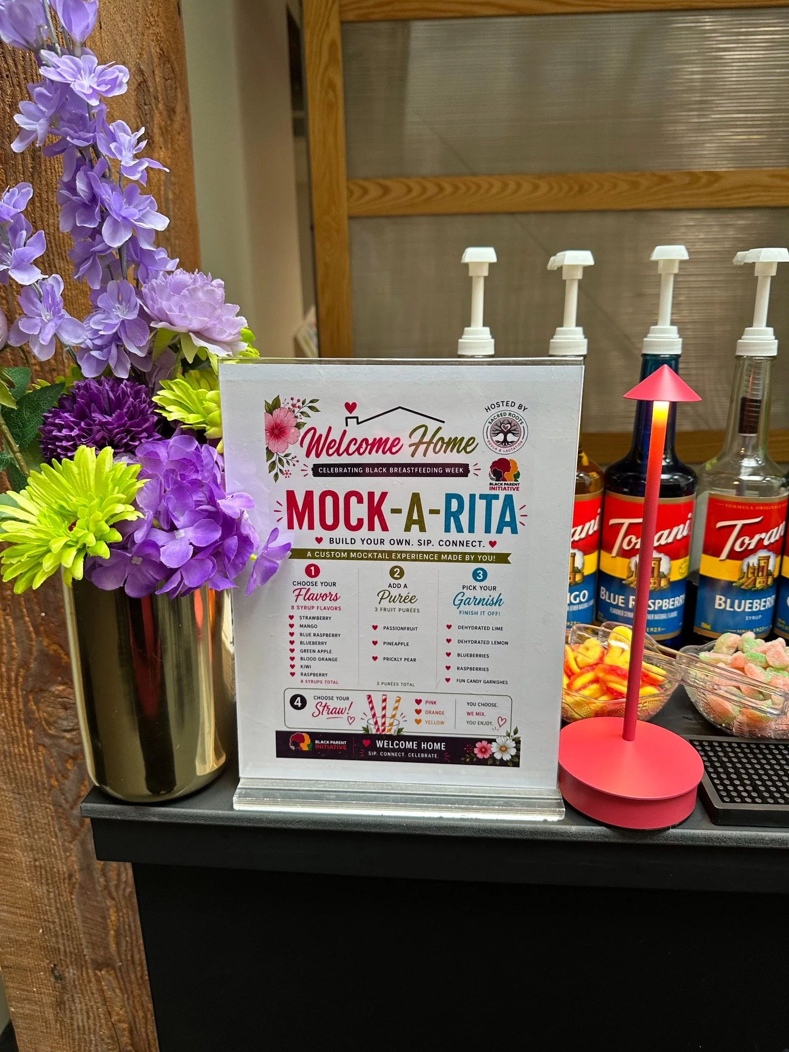 Mock-A-Rita custom mocktail menu beside purple flowers and colorful syrup bottles