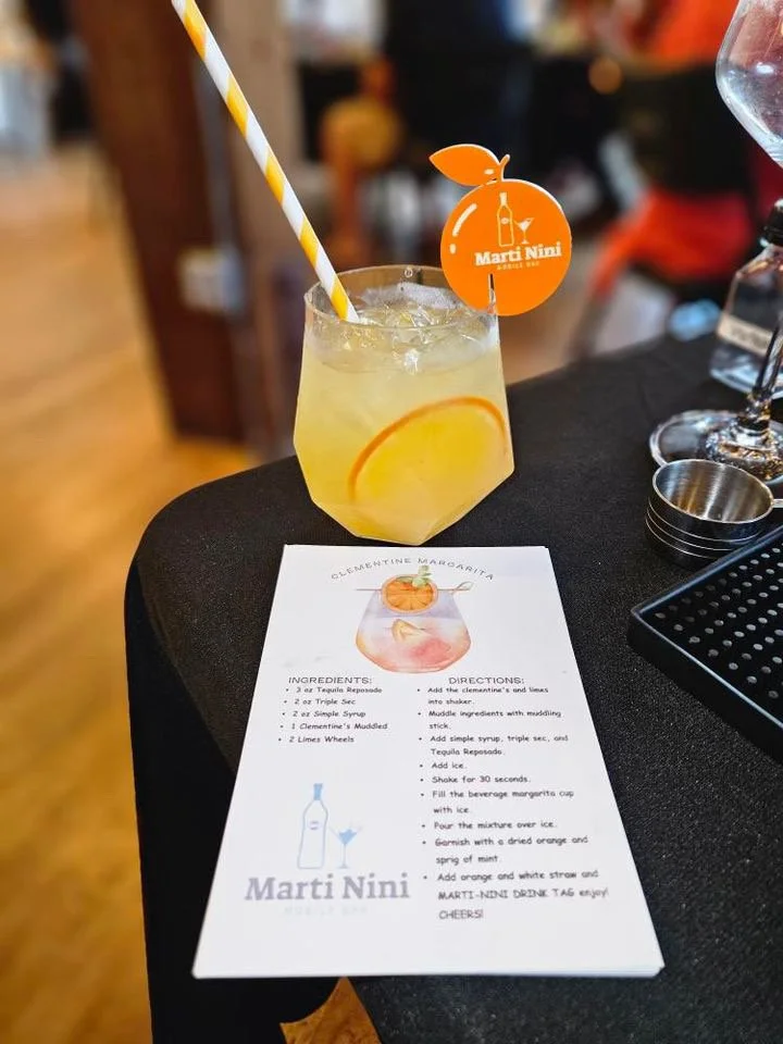 Yellow-orange cocktail beside a Marti NiNi recipe card and branded garnish