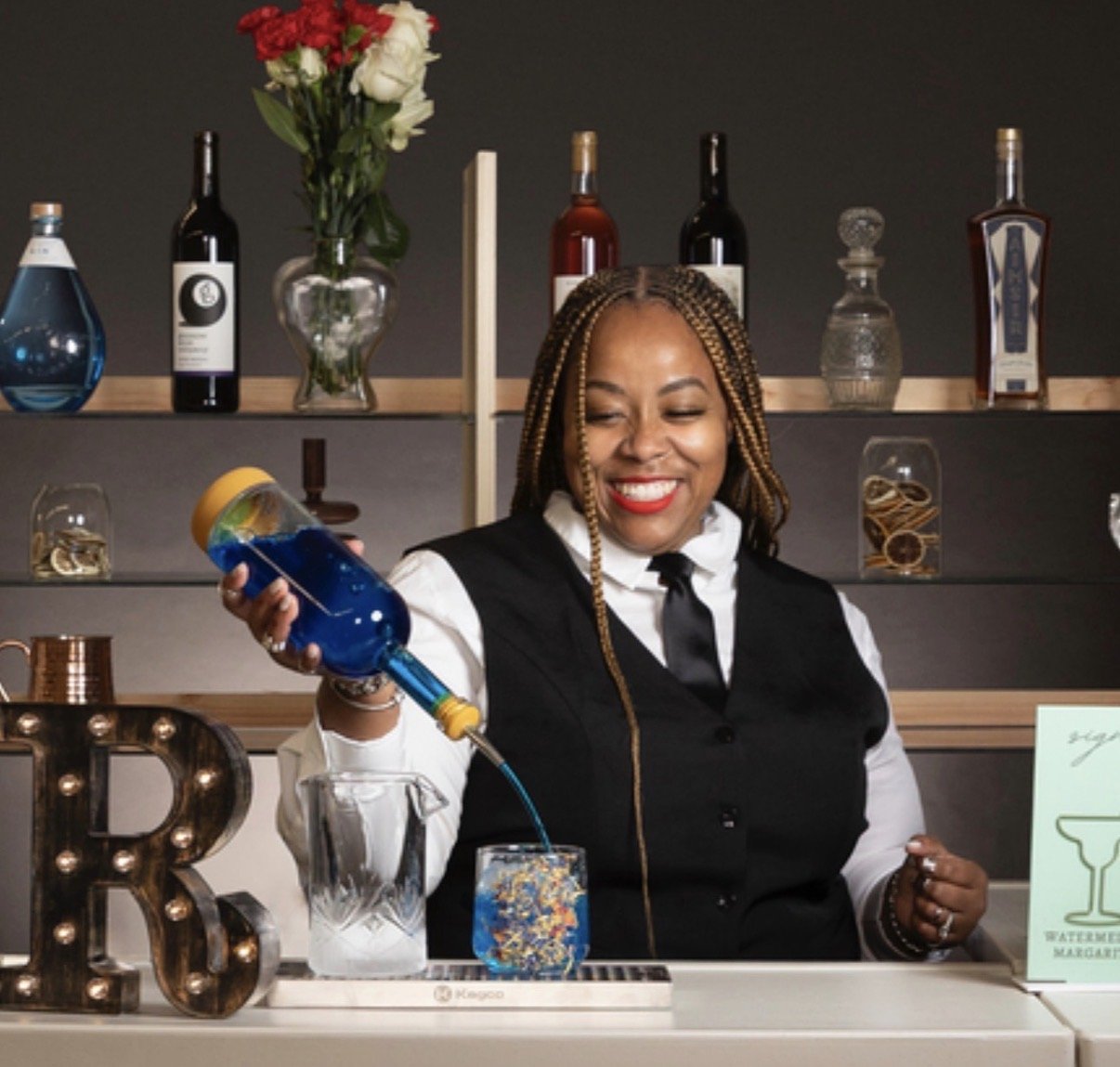NiNi smiles while pouring a bright blue cocktail behind the bar