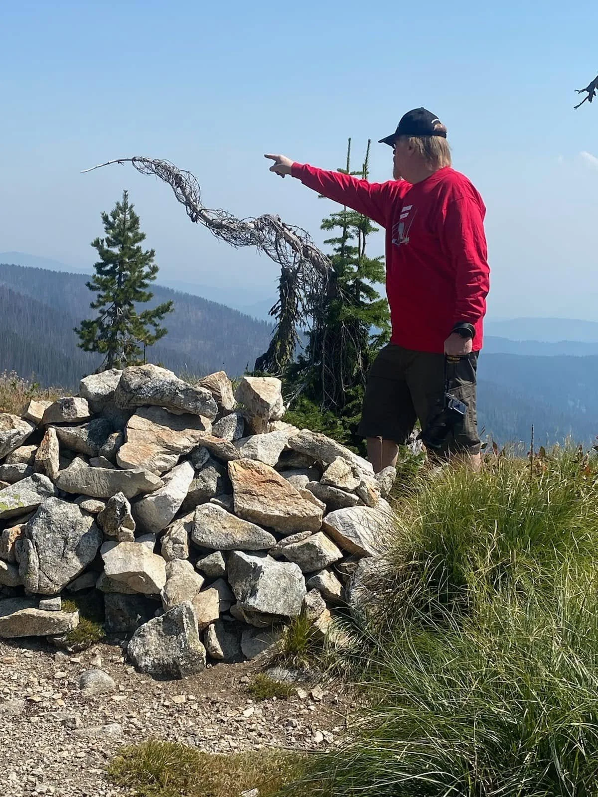 Scott Gray, wearing a red shirt and cap, points across a rocky mountain landscape.