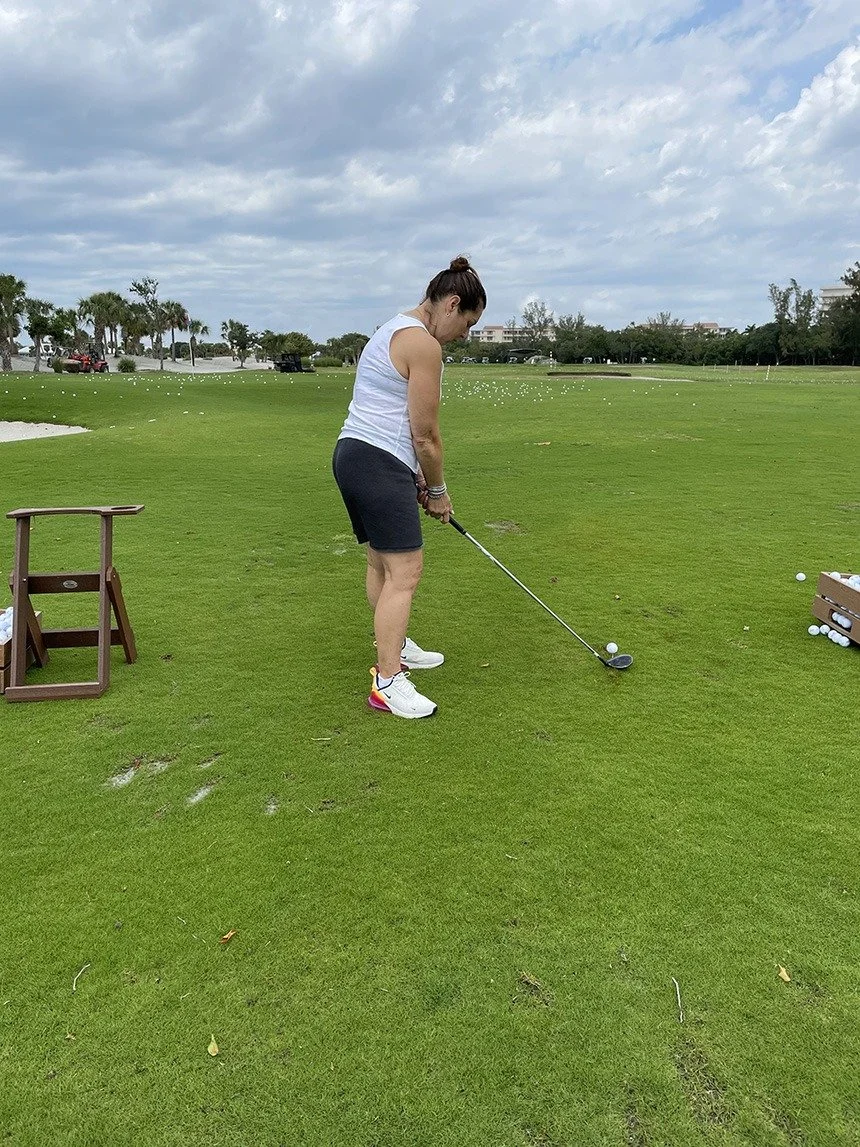 A woman in golf attire mid-swing on a driving range, palm trees and a resort building in the background