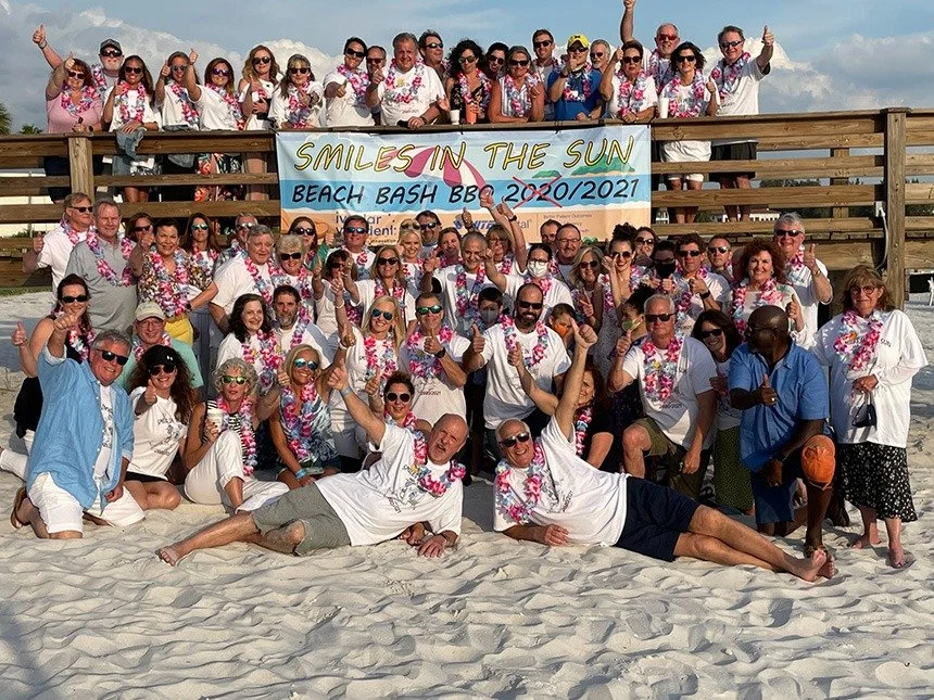 A large group of attendees posing together on a beach boardwalk beside a banner reading Smiles in the Sun Beach Bash BBQ 2020/2021
