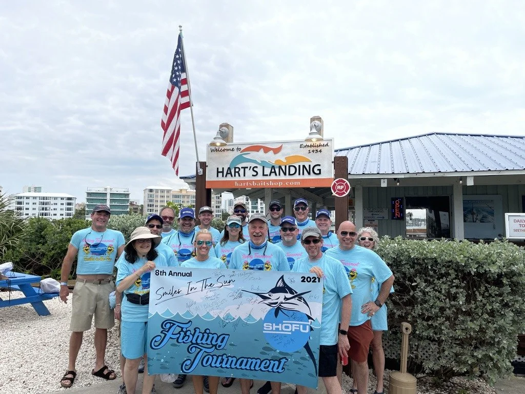 A group in matching blue shirts posing with a banner reading 8th Annual Smiles in the Sun Fishing Tournament 2021 at Hart's Landing