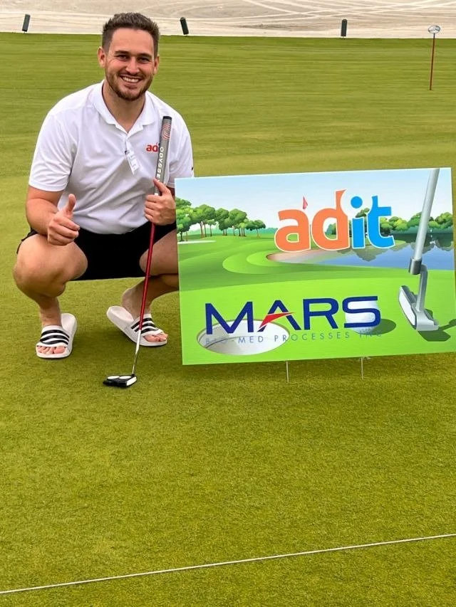 A man kneeling on a golf green next to a tee sign for sponsors adit and MARS Bio-Med Processes