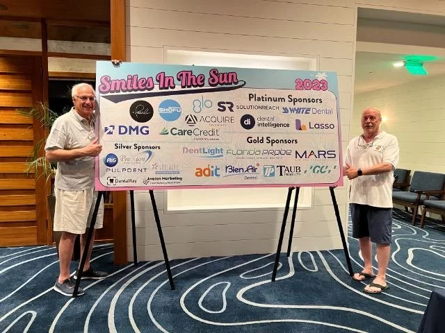 Two men posing beside a Smiles in the Sun 2023 sponsor recognition board listing platinum, gold and silver sponsors