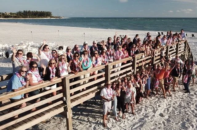 A large group of attendees wearing floral leis, gathered on a beach boardwalk waving at the camera