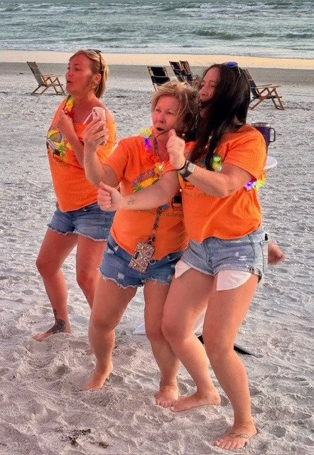 Three women in matching orange event t-shirts dancing together on the beach in the evening light