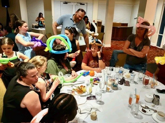 Attendees at a banquet dinner wearing balloon-animal party hats and laughing together at the table