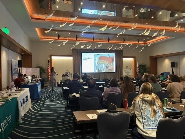 Attendees seated at tables in a hotel conference room watching a presenter speak beside a projection screen, with sponsor tables along the left wall