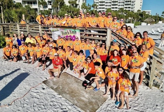 A large group in matching orange shirts and leis posing together on a beach boardwalk, with a Beach Bash banner