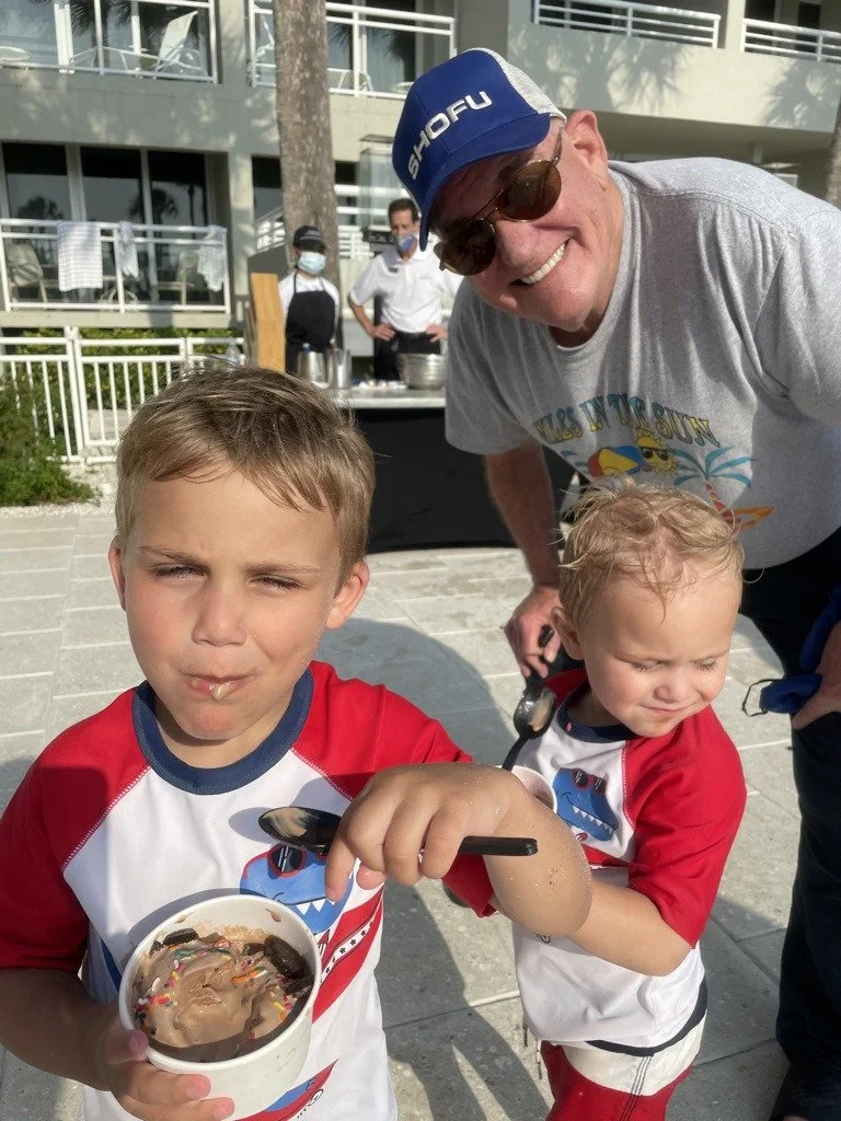 Two young children eating ice cream treats poolside while a man in a Smiles in the Sun t-shirt looks on smiling