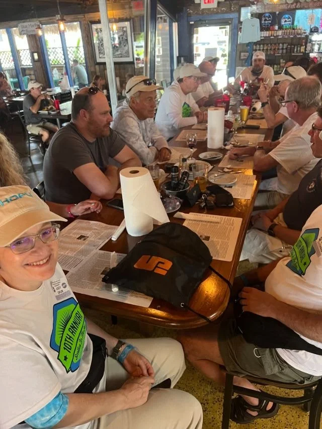 A group of attendees seated around a table at a casual waterfront restaurant, one wearing a shirt marking the 10th Annual tournament