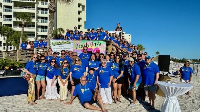 A large group of attendees in matching blue Smiles in the Sun t-shirts posing together on the beach in front of a Beach Bash banner, with resort buildings behind them