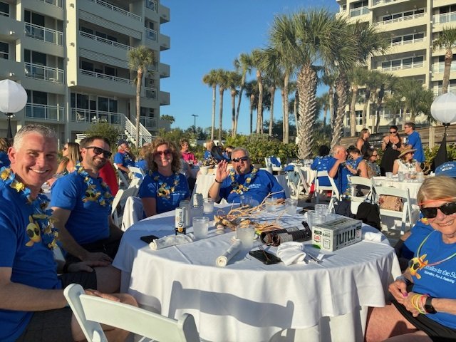 Attendees in blue event t-shirts and floral leis seated around a white-linen table outdoors, smiling at the camera with palm trees and the resort behind them