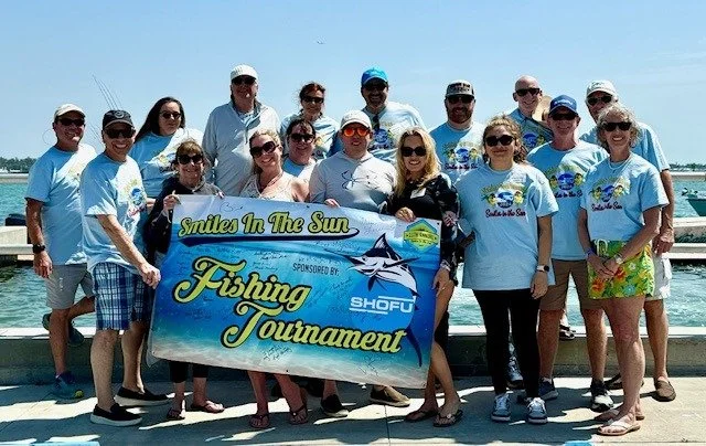 A group in matching light blue shirts posing on a dock with a Smiles in the Sun Fishing Tournament banner