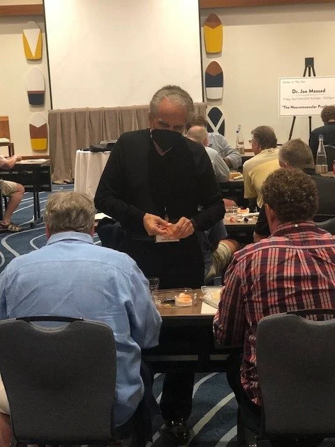 A presenter demonstrating dental models to seated attendees at a hands-on workshop table, with a sign for Dr. Joe Massad's Neuromuscular Program session