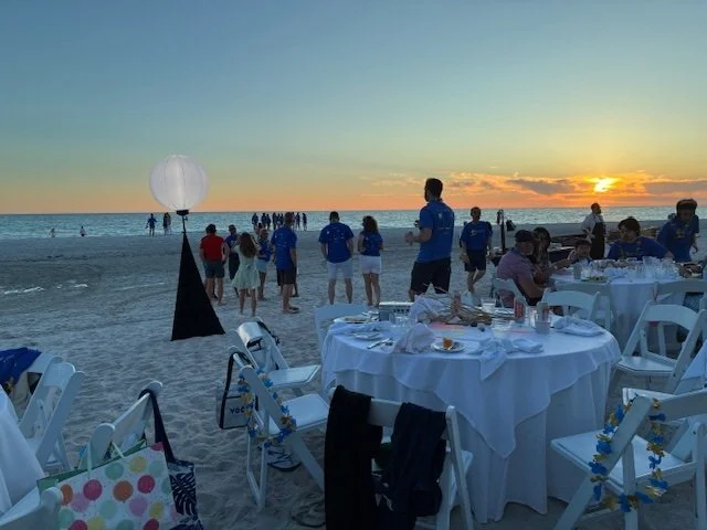 Round banquet tables set with white linens on the sand at sunset, with guests gathered nearby as the sun sets over the Gulf