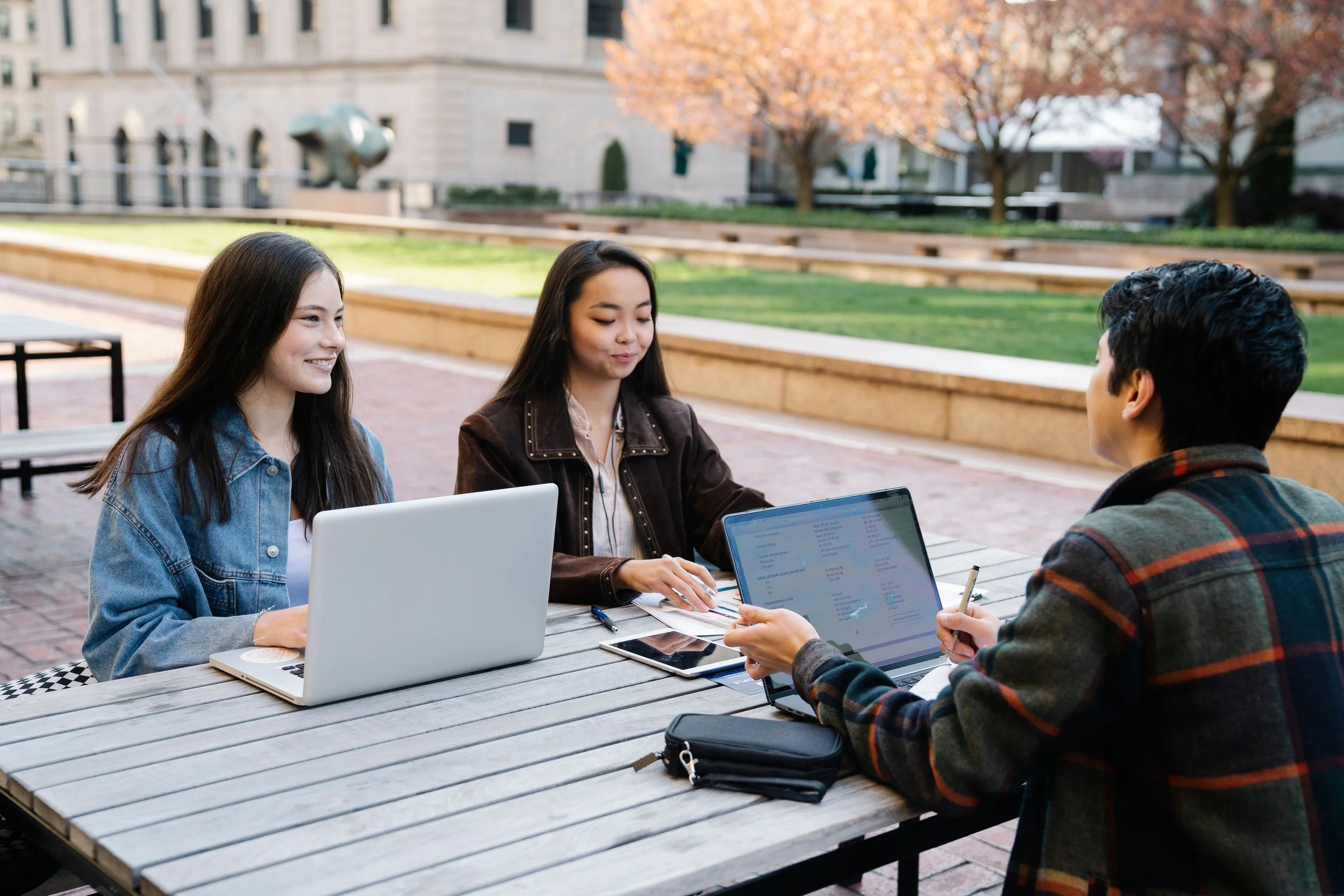 Three adults studying together outdoors with laptops and notebooks.