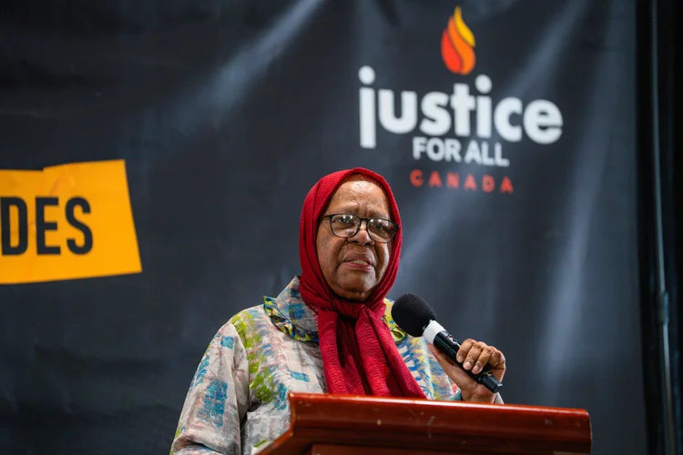 A speaker at the podium in front of the Justice for All Canada banner.