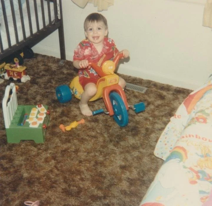 A toddler grinning on a red and yellow tricycle in a carpeted living room