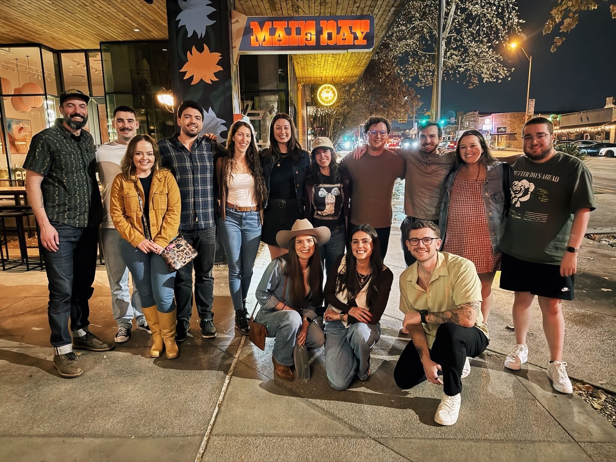 A group of fourteen colleagues smiling for a photo outside a restaurant at night