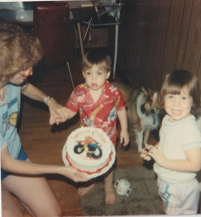 Two small kids and a collie gathered around a birthday cake