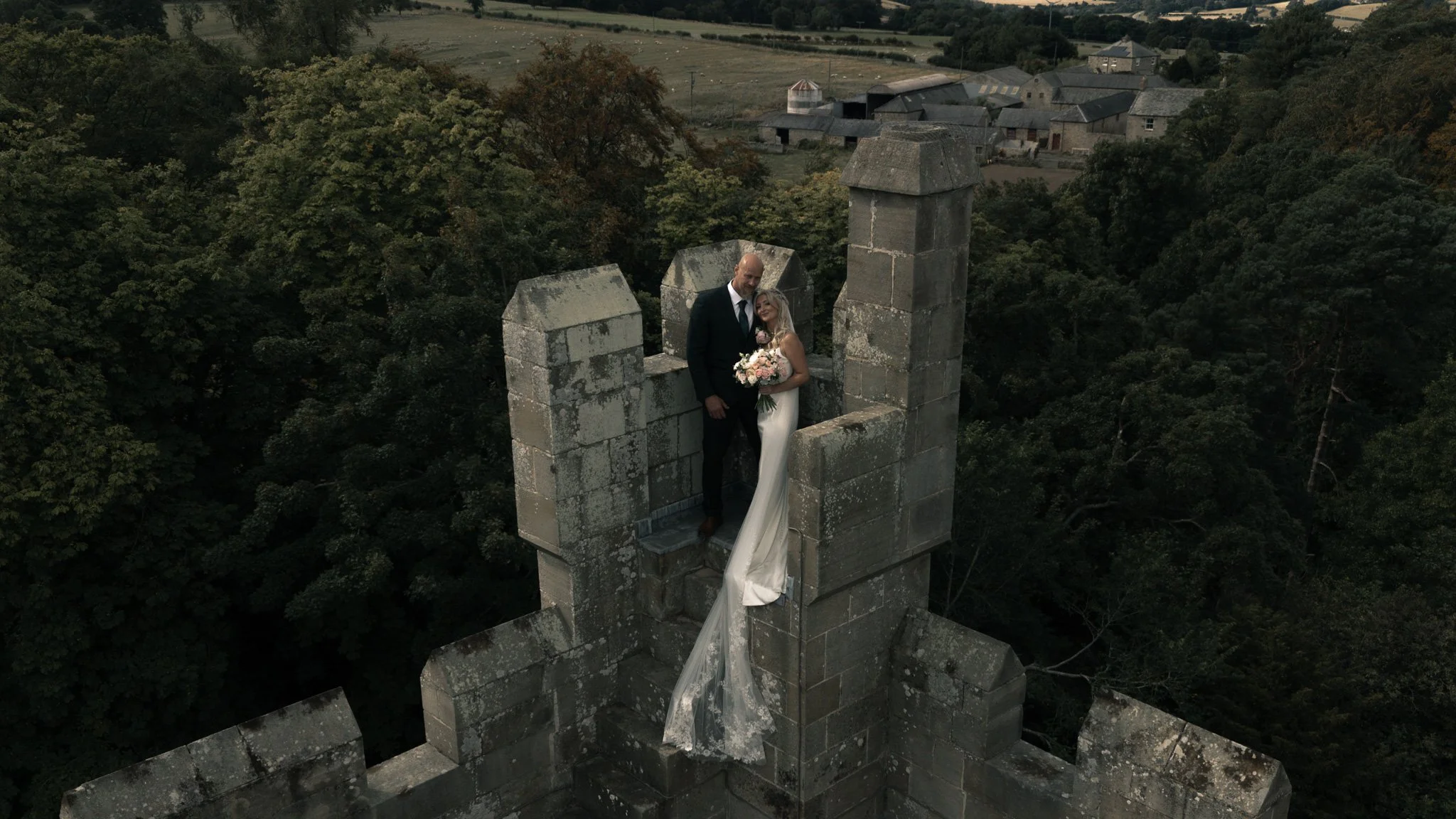 Bride and groom standing together among castle battlements, with a long veil flowing down the stonework.