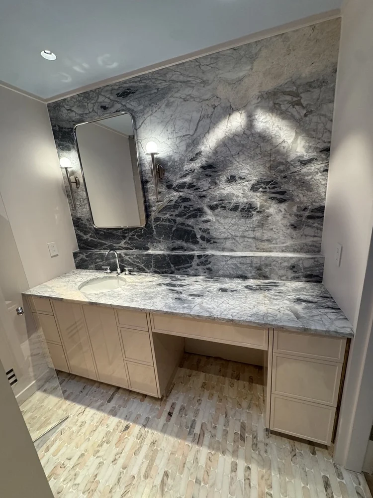 Gray-and-black veined vanity counter and feature wall with globe sconces and warm cabinetry.