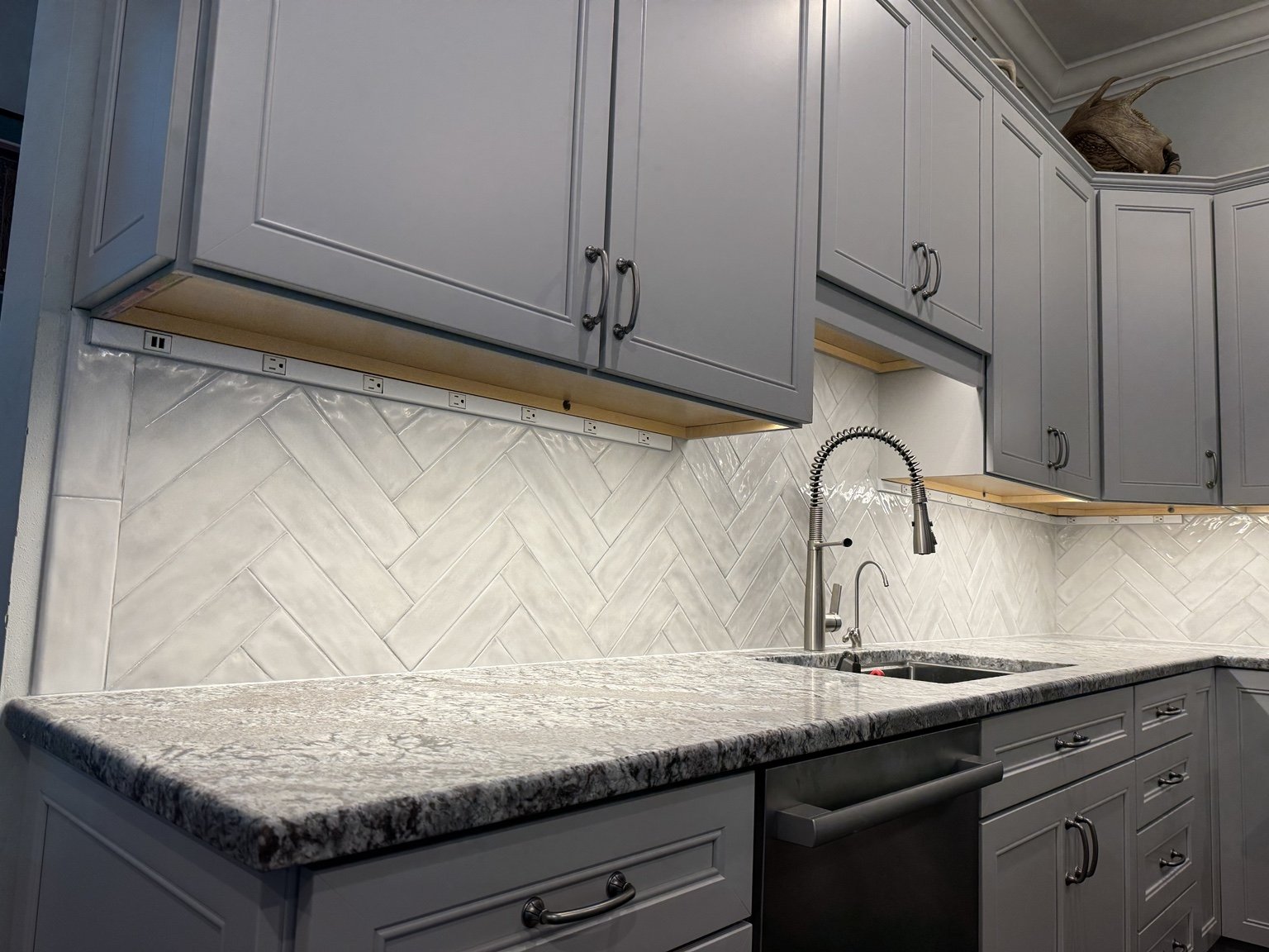 White herringbone backsplash beneath gray cabinets, with under-cabinet lighting and a spring-coil kitchen faucet.