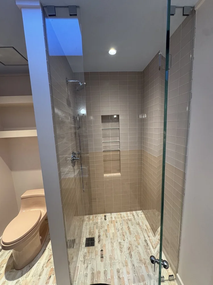 Glass-enclosed taupe tile shower with chrome fixtures, a tall shelving niche, and mosaic floor.