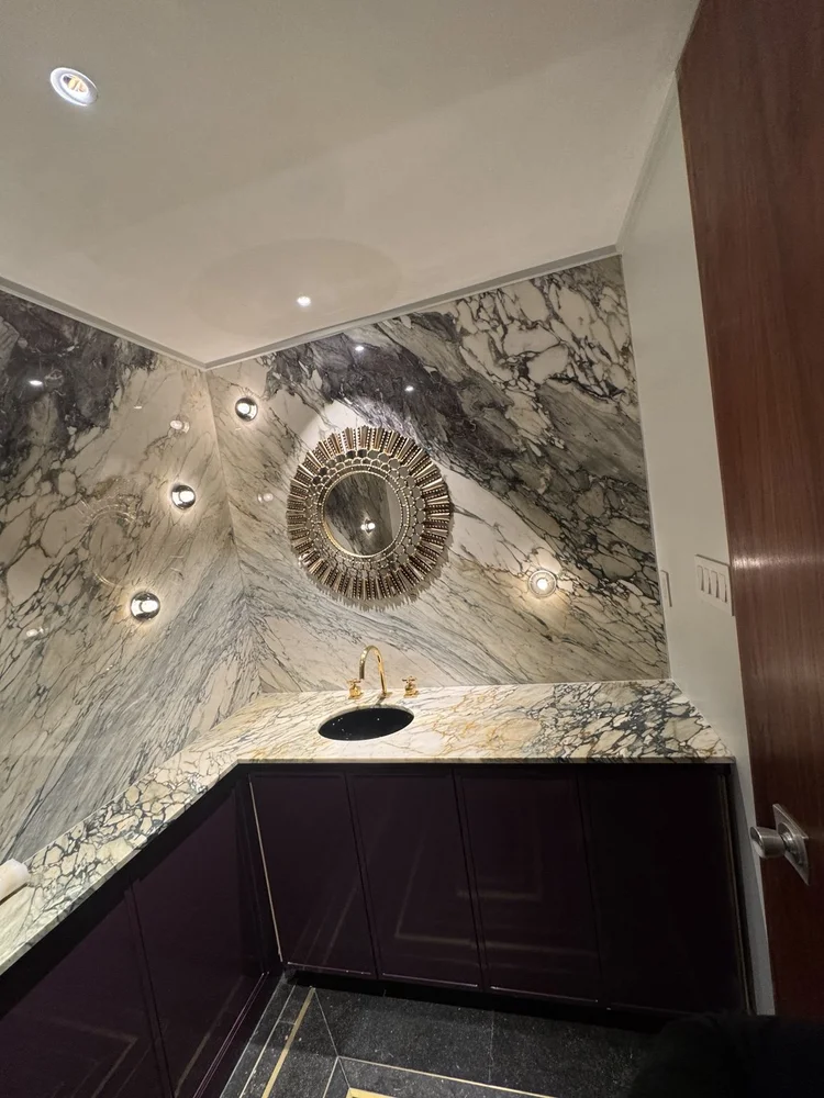 Dark veined vanity with a circular mirror, gold faucet, and black floor with brass inlay.