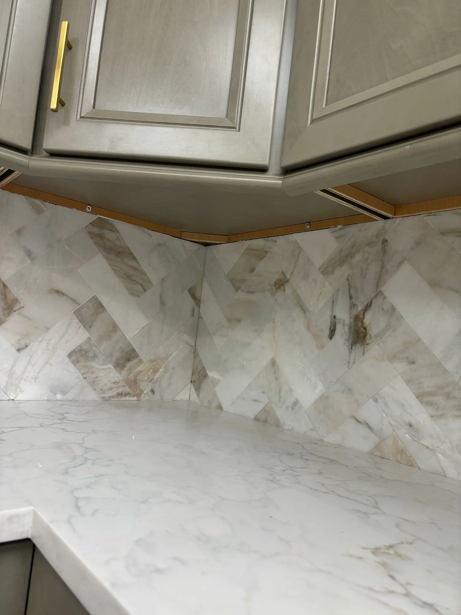 Close-up of veined herringbone backsplash tile meeting a light counter beneath gray cabinetry with gold hardware.