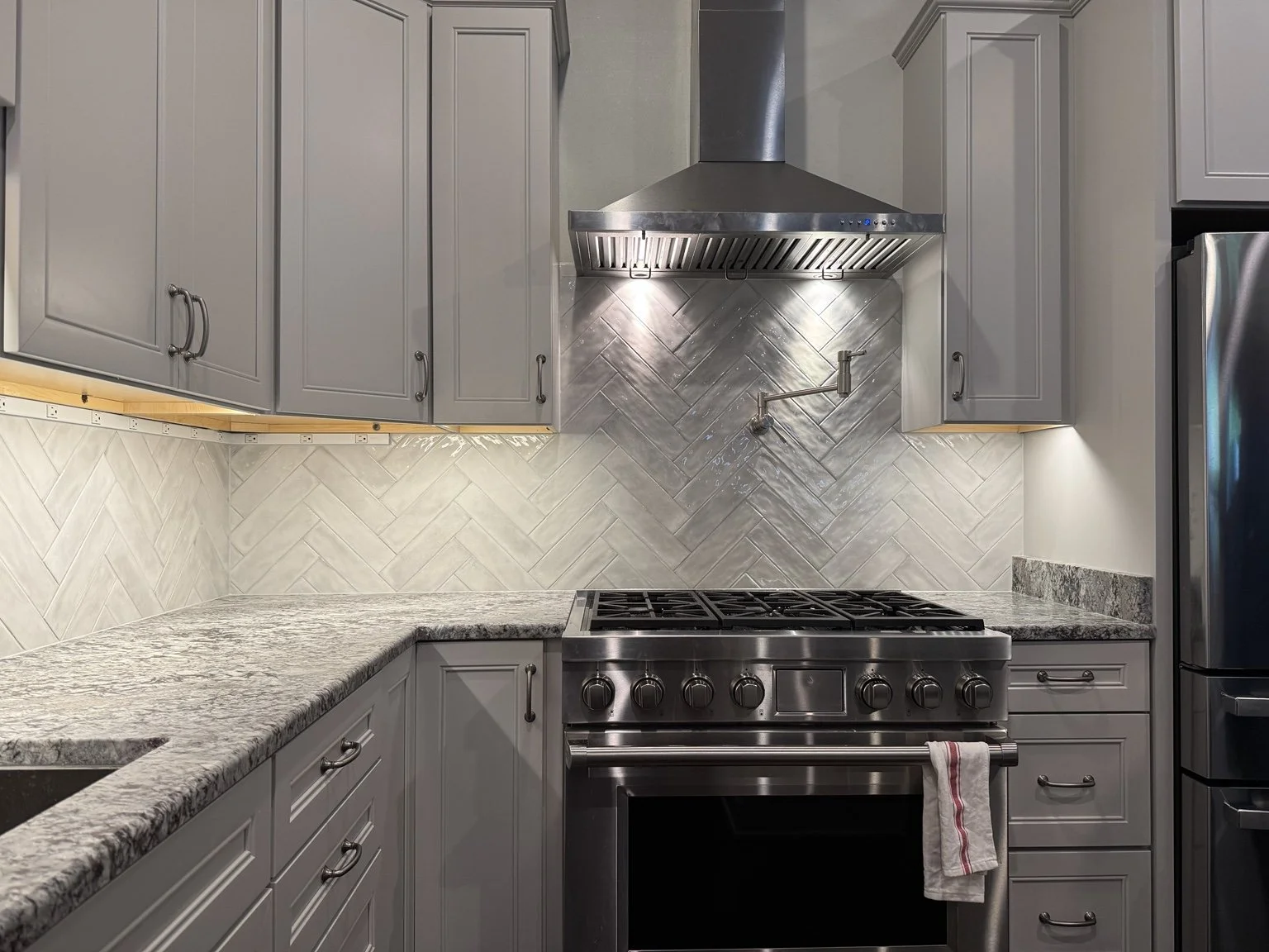 Gray kitchen cabinetry framing a stainless range and hood, with an illuminated white herringbone backsplash.