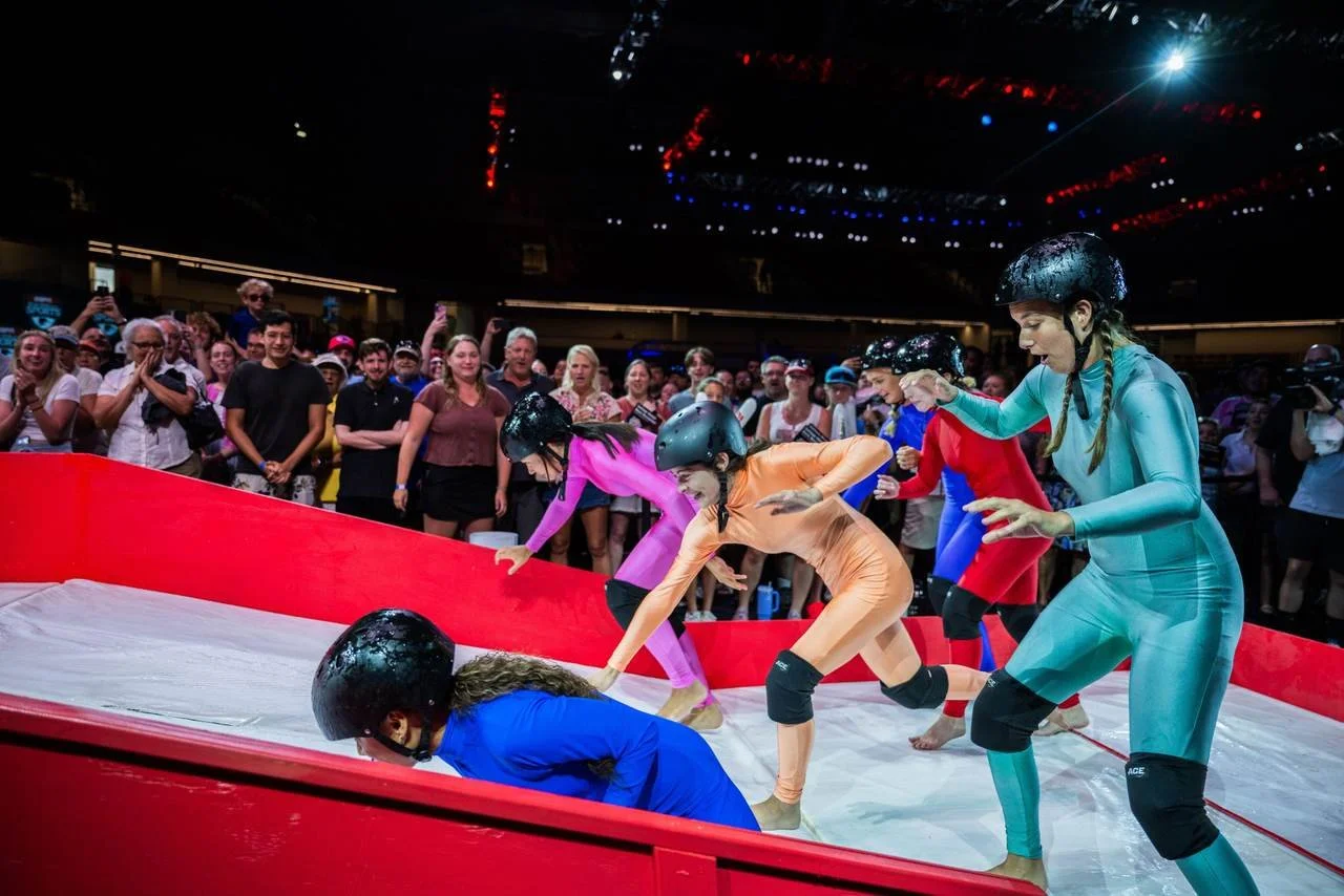 Competitors in helmets and coloured bodysuits scramble up a slippery inflatable ramp in front of a cheering crowd at a Slippery Stairs event.